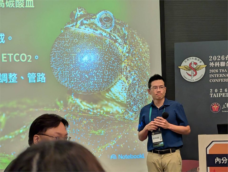 梁宗榮醫師在 2026 台灣外科醫學會聯合學術年會的講台上手持麥克風報告，身後投影片是一張蛙類的影像
