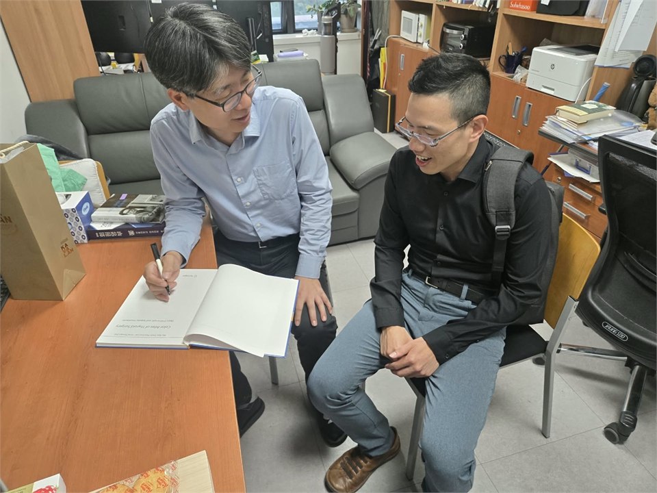 辦公室裡的照片：一位穿淺藍色襯衫、戴眼鏡的男醫師站在書桌旁，彎身在攤開的書上簽名，扉頁朝著他、對鏡頭是上下顛倒的；另一位戴眼鏡、穿深色襯衫背著後背包的男醫師坐在旁邊的椅子上笑著看他。桌上另有一個紙袋，背後是沙發與書櫃