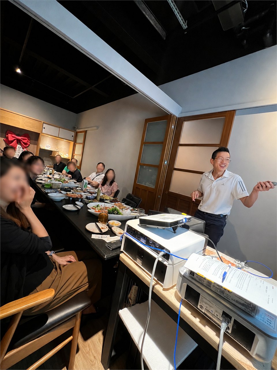 演講當天的照片：餐廳包廂裡，講者站在右側的投影設備旁講解，左側長桌坐著十餘位聽眾，桌上擺著餐點；聽眾臉部均已模糊處理