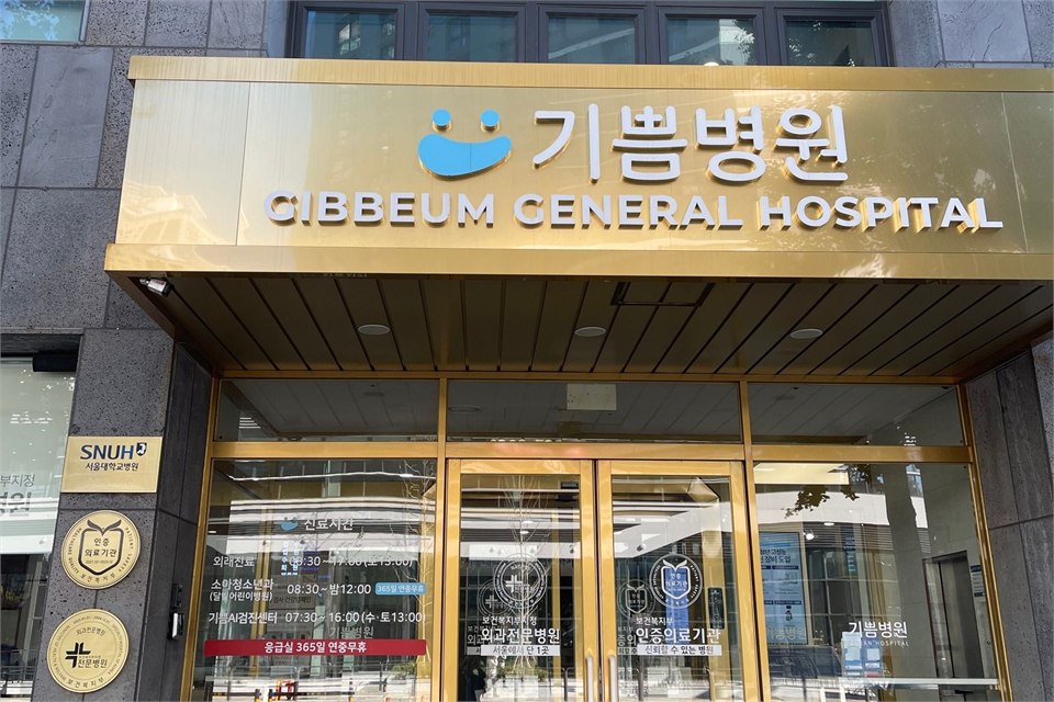 醫院正門的照片：灰色石材外牆的多層建築，一樓是金色框的玻璃門，門楣上方的金色招牌寫著韓文「기쁨병원」與英文 GIBBEUM GENERAL HOSPITAL