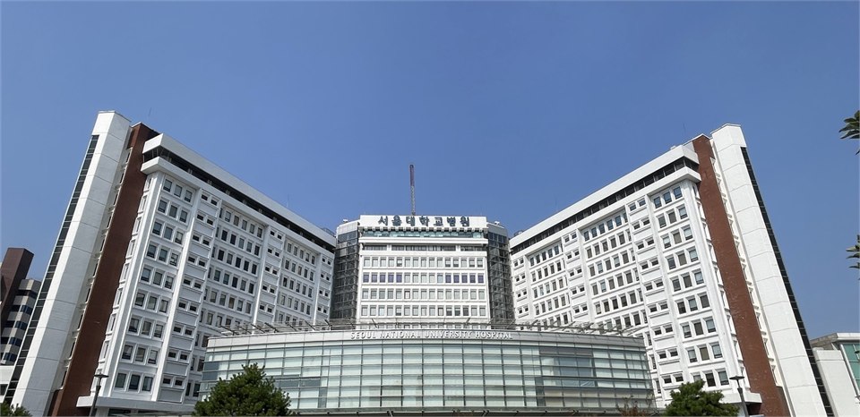 首爾大學醫院主體建築的外觀照片：中央是弧形玻璃帷幕的門診大樓，正面寫著 SEOUL NATIONAL UNIVERSITY HOSPITAL，上方塔樓掛著韓文院名 서울대학교병원，左右各有一棟白色的住院大樓向前伸出