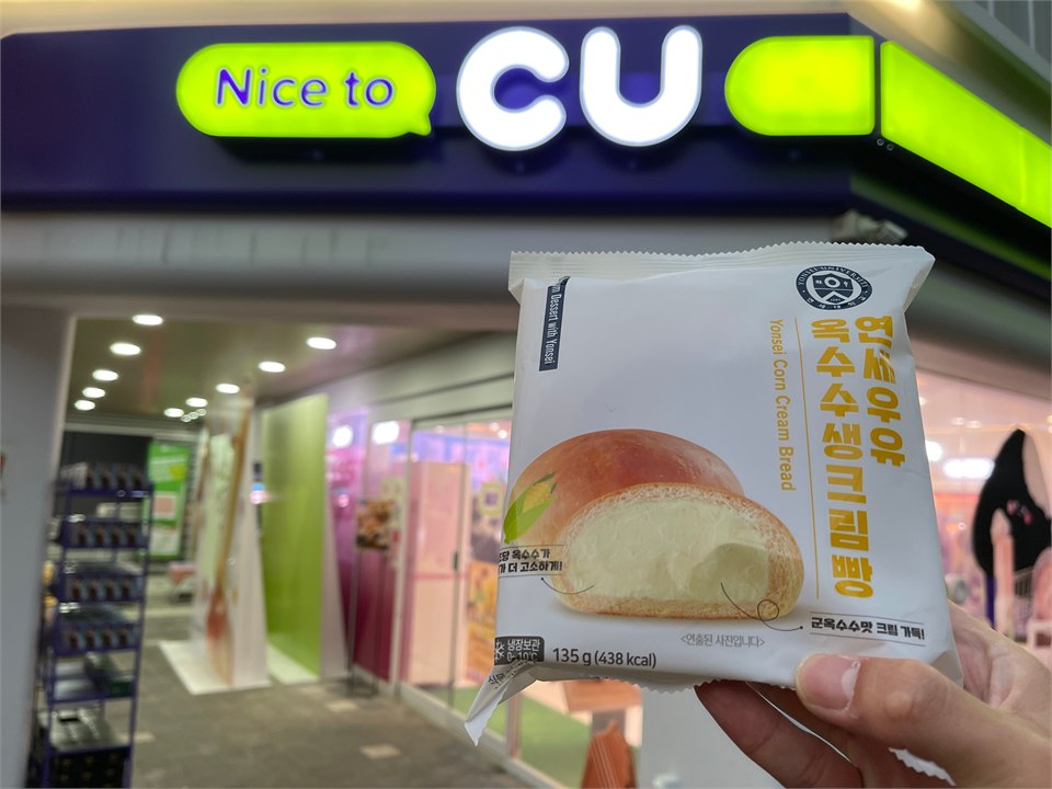 一隻手舉著一包生乳包，背景是便利商店 CU 的招牌與店門。包裝上有延世大學的校徽與韓文「연세우유 옥수수생크림빵」，英文小字 Yonsei Corn Cream Bread